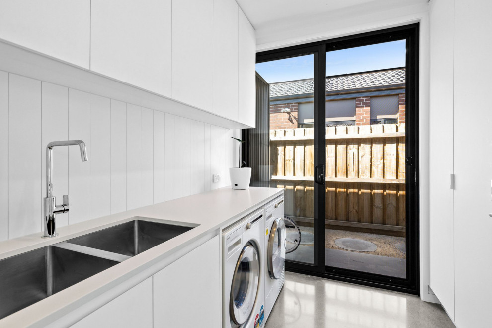 This is an example of a mid-sized galley dedicated laundry room in Geelong with a double-bowl sink, flat-panel cabinets, white cabinets, quartz benchtops, white splashback, ceramic splashback, white walls, concrete floors, a side-by-side washer and dryer and grey floor.