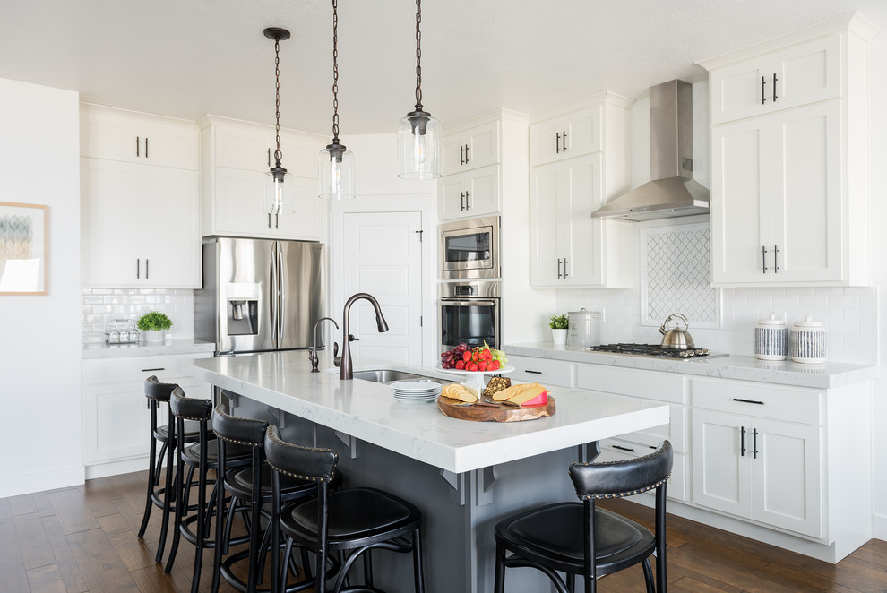 Inspiration for a transitional l-shaped kitchen in Salt Lake City with an undermount sink, shaker cabinets, white cabinets, white splashback, subway tile splashback, stainless steel appliances, dark hardwood floors, with island, brown floor and white benchtop.