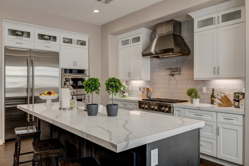This is an example of a transitional l-shaped kitchen in Las Vegas with a farmhouse sink, shaker cabinets, white cabinets, grey splashback, stainless steel appliances, dark hardwood floors, with island, brown floor and white benchtop.