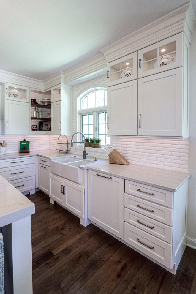 Inspiration for a large transitional u-shaped eat-in kitchen in Cleveland with a farmhouse sink, flat-panel cabinets, white cabinets, quartz benchtops, white splashback, ceramic splashback, white appliances, dark hardwood floors, with island, brown floor and grey benchtop.