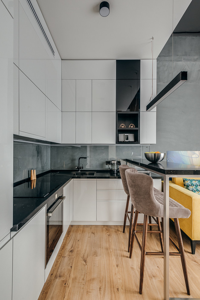 Inspiration for an industrial l-shaped open plan kitchen in Moscow with flat-panel cabinets, grey splashback, stone slab splashback, black appliances, medium hardwood floors, with island, brown floor and black benchtop.