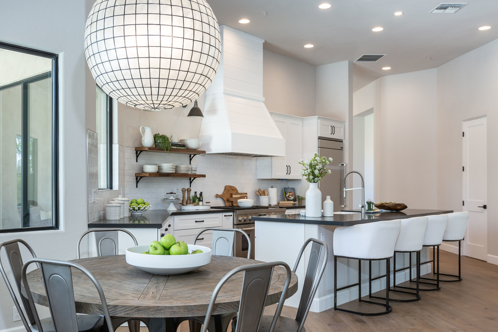 Transitional l-shaped medium tone wood floor and brown floor kitchen photo in Phoenix with shaker cabinets, white cabinets, white backsplash, stainless steel appliances, an island and black countertops