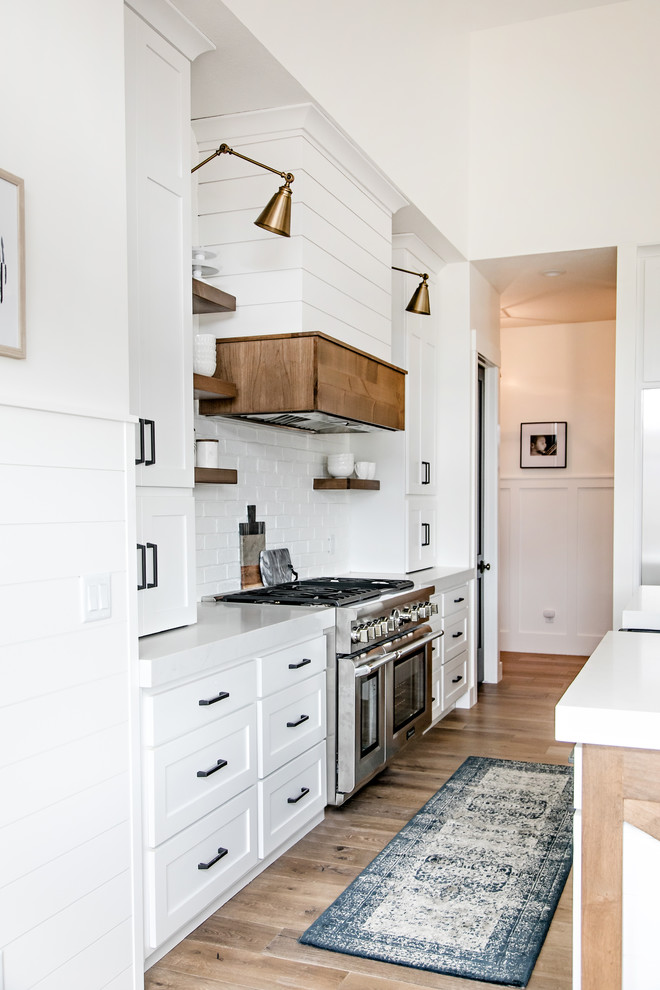 Mid-sized country l-shaped eat-in kitchen in Salt Lake City with a farmhouse sink, shaker cabinets, white cabinets, quartz benchtops, white splashback, subway tile splashback, stainless steel appliances, medium hardwood floors, with island, brown floor and white benchtop.