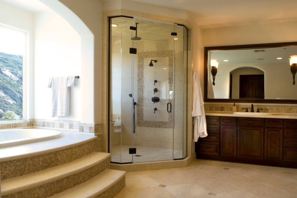 Design ideas for a mediterranean bathroom in Los Angeles with a drop-in sink, raised-panel cabinets, dark wood cabinets, limestone benchtops, a drop-in tub, a corner shower, a one-piece toilet, beige tile and stone tile.