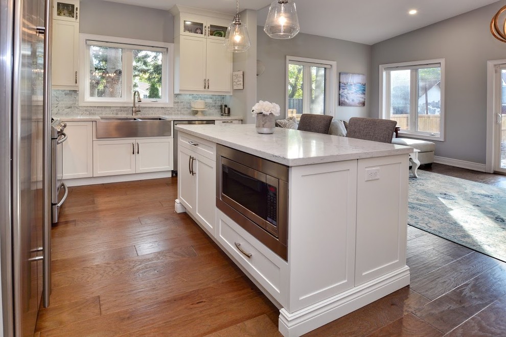 Example of a mid-sized transitional l-shaped medium tone wood floor and brown floor eat-in kitchen design in Toronto with a farmhouse sink, shaker cabinets, white cabinets, quartz countertops, gray backsplash, marble backsplash, stainless steel appliances, an island and white countertops