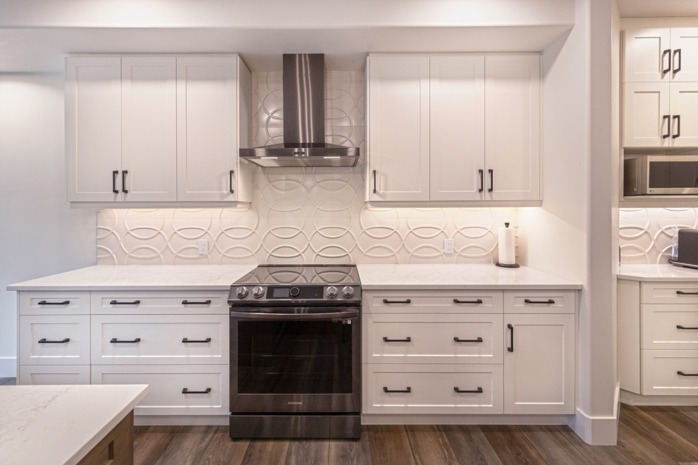 Mid-sized traditional l-shaped eat-in kitchen in Edmonton with a double-bowl sink, recessed-panel cabinets, white cabinets, quartz benchtops, white splashback, ceramic splashback, stainless steel appliances, vinyl floors, with island, brown floor and multi-coloured benchtop.