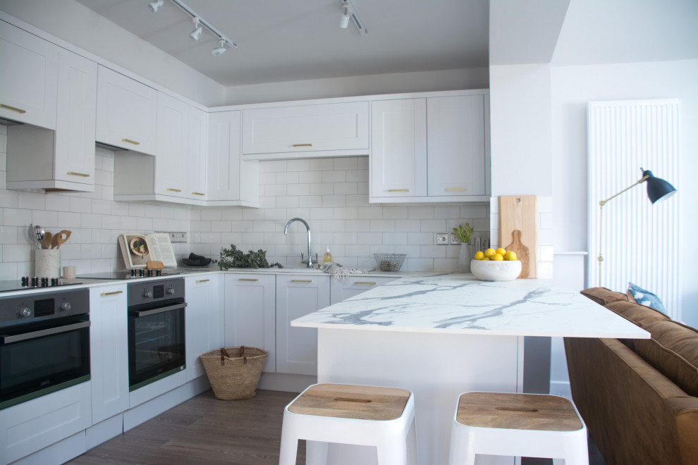 Inspiration for a mid-sized transitional u-shaped open plan kitchen in Cambridgeshire with a double-bowl sink, shaker cabinets, white cabinets, laminate benchtops, white splashback, laminate floors, a peninsula, brown floor, subway tile splashback, stainless steel appliances and grey benchtop.
