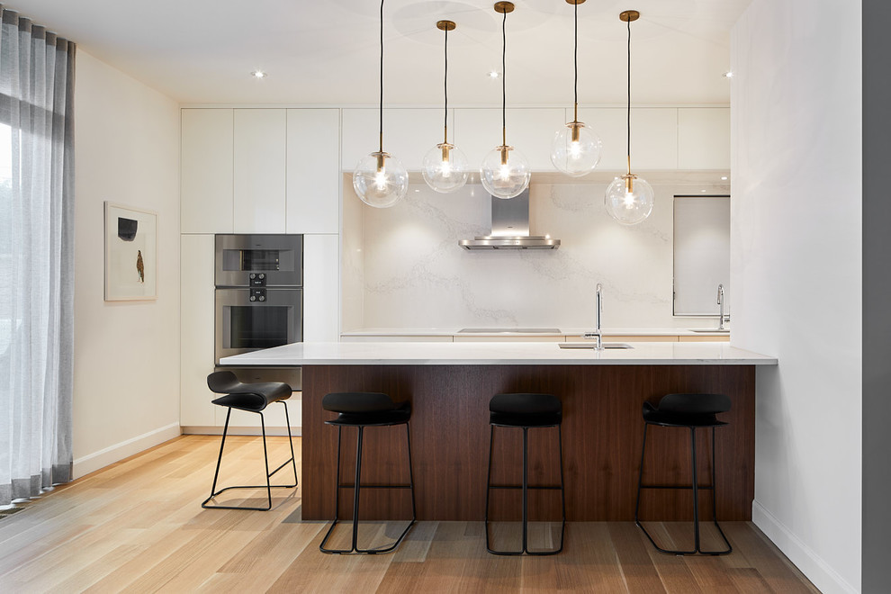 Inspiration for a contemporary galley kitchen in Ottawa with an undermount sink, flat-panel cabinets, white cabinets, white splashback, marble splashback, black appliances, light hardwood floors, a peninsula, beige floor and white benchtop.