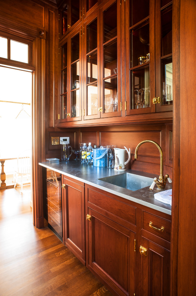 Inspiration for a small traditional galley wet bar in Portland Maine with an integrated sink, shaker cabinets, dark wood cabinets, stainless steel benchtops, brown splashback, timber splashback, medium hardwood floors and brown floor.