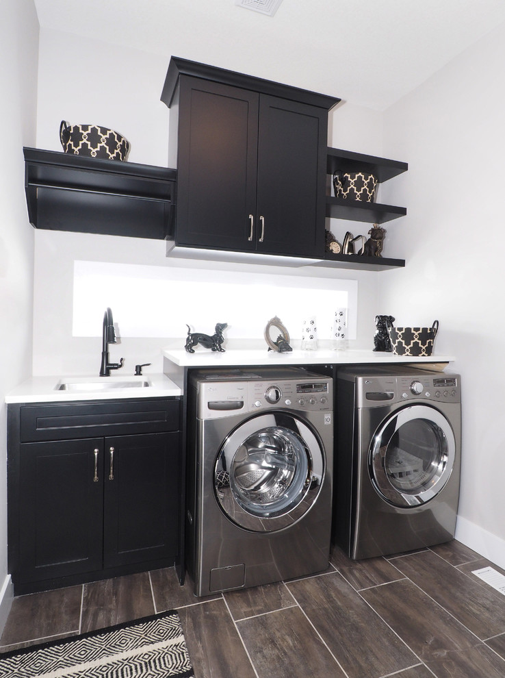 Design ideas for a mid-sized transitional single-wall dedicated laundry room in Toronto with an undermount sink, shaker cabinets, black cabinets, white walls, a side-by-side washer and dryer, brown floor and white benchtop.