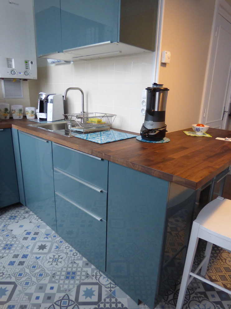 Small contemporary l-shaped eat-in kitchen in Lyon with a single-bowl sink, beaded inset cabinets, blue cabinets, wood benchtops, beige splashback, ceramic splashback, stainless steel appliances, cement tiles, no island, blue floor and brown benchtop.
