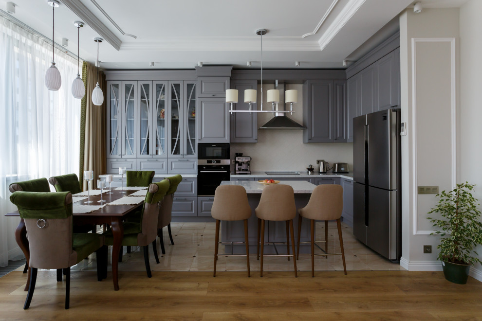 This is an example of a mid-sized transitional l-shaped open plan kitchen in Moscow with an undermount sink, raised-panel cabinets, grey cabinets, quartz benchtops, white splashback, ceramic splashback, stainless steel appliances, ceramic floors, with island, beige floor and white benchtop.