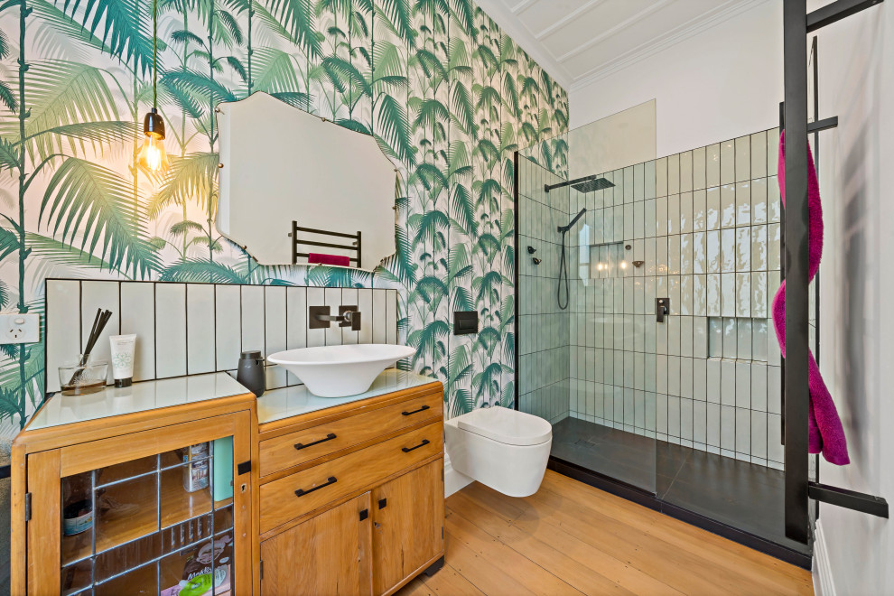 This is an example of a large eclectic kids bathroom in Auckland with furniture-like cabinets, brown cabinets, an open shower, a wall-mount toilet, green tile, subway tile, green walls, medium hardwood floors, a vessel sink, glass benchtops, brown floor, an open shower, green benchtops, a single vanity, a freestanding vanity, exposed beam and wallpaper.
