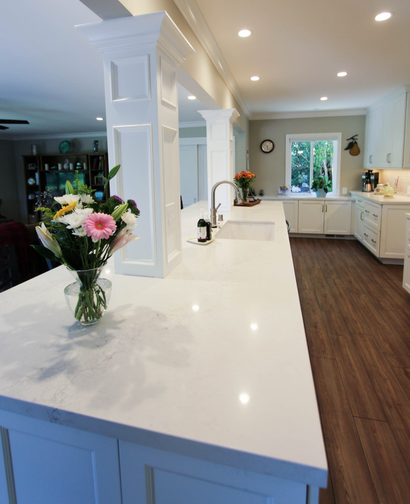 This is an example of a large classic galley open plan kitchen in San Francisco with a submerged sink, recessed-panel cabinets, white cabinets, engineered stone countertops, grey splashback, metro tiled splashback, stainless steel appliances, vinyl flooring, an island, brown floors and white worktops.