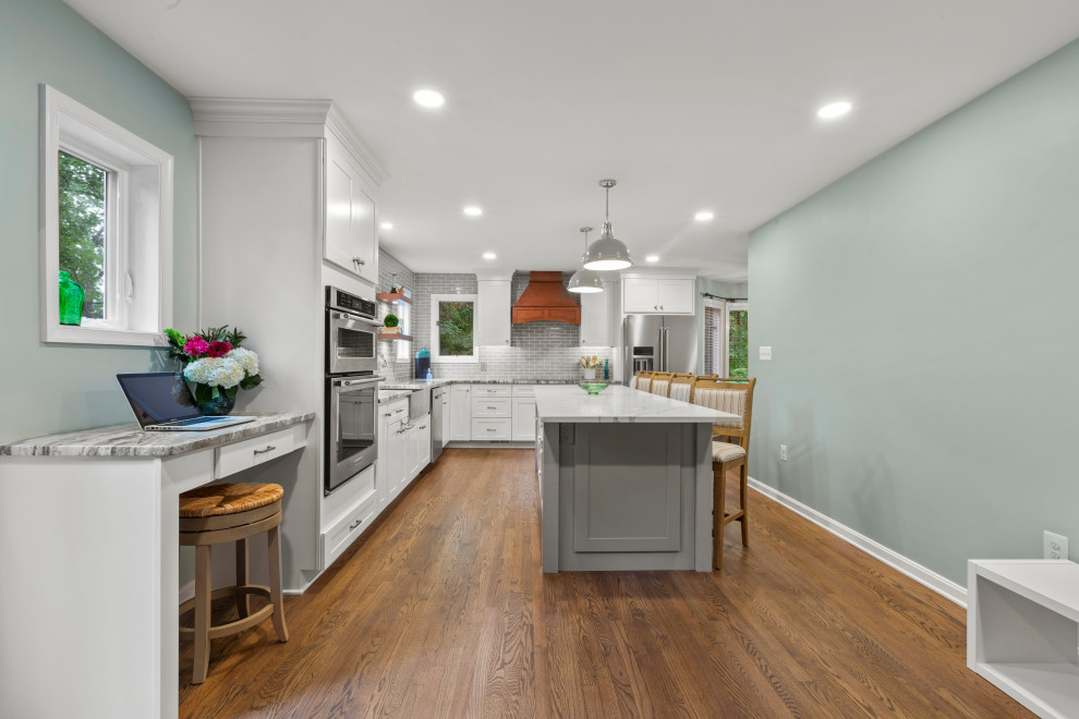 Inspiration for a traditional eat-in kitchen in DC Metro with a farmhouse sink, recessed-panel cabinets, white cabinets, granite benchtops, grey splashback, ceramic splashback, stainless steel appliances, light hardwood floors, with island, brown floor and white benchtop.