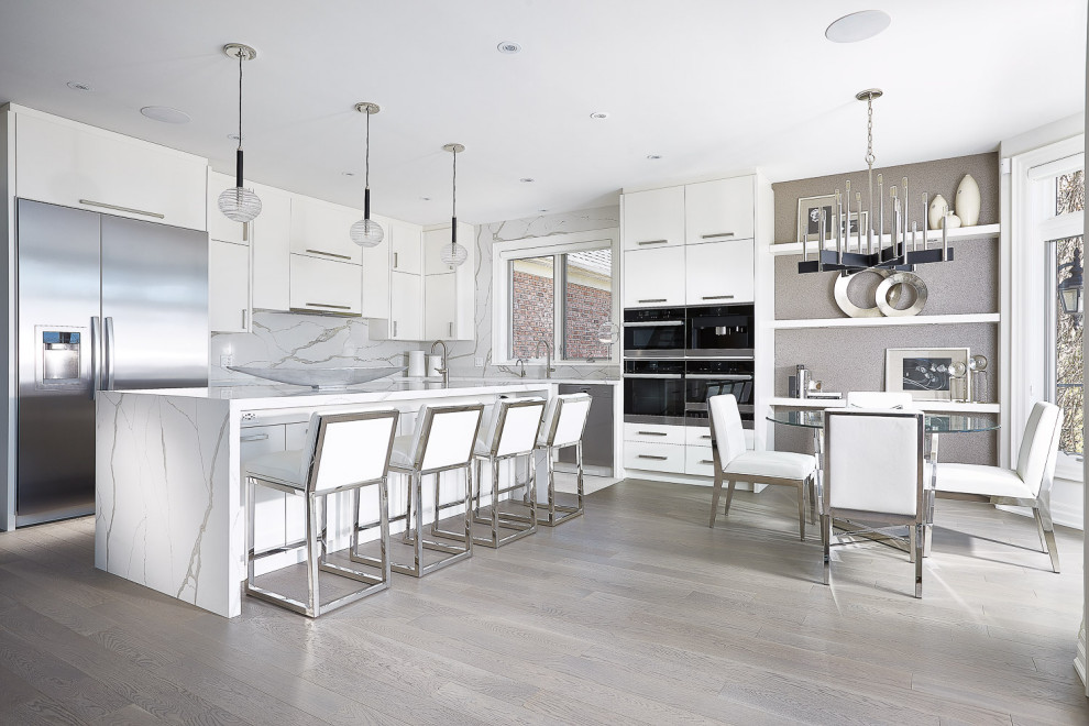 This is an example of a large contemporary l-shaped eat-in kitchen in Toronto with flat-panel cabinets, white cabinets, white splashback, stainless steel appliances, light hardwood floors, with island, white benchtop, stone slab splashback and beige floor.