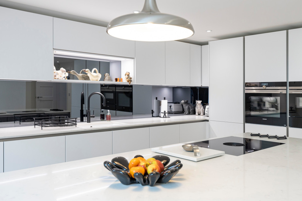 Inspiration for a mid-sized contemporary galley eat-in kitchen in Hertfordshire with a single-bowl sink, flat-panel cabinets, white cabinets, quartzite benchtops, grey splashback, mirror splashback, black appliances, light hardwood floors, with island, brown floor, white benchtop and recessed.