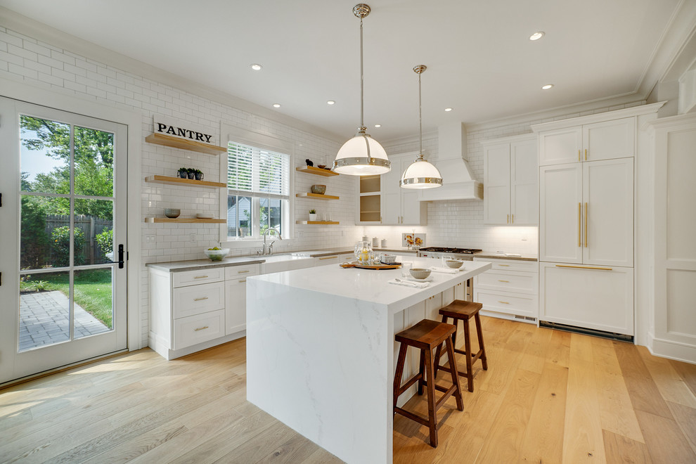 Example of a cottage l-shaped light wood floor and beige floor kitchen design in Philadelphia with a farmhouse sink, shaker cabinets, white cabinets, white backsplash, subway tile backsplash, paneled appliances, an island and gray countertops