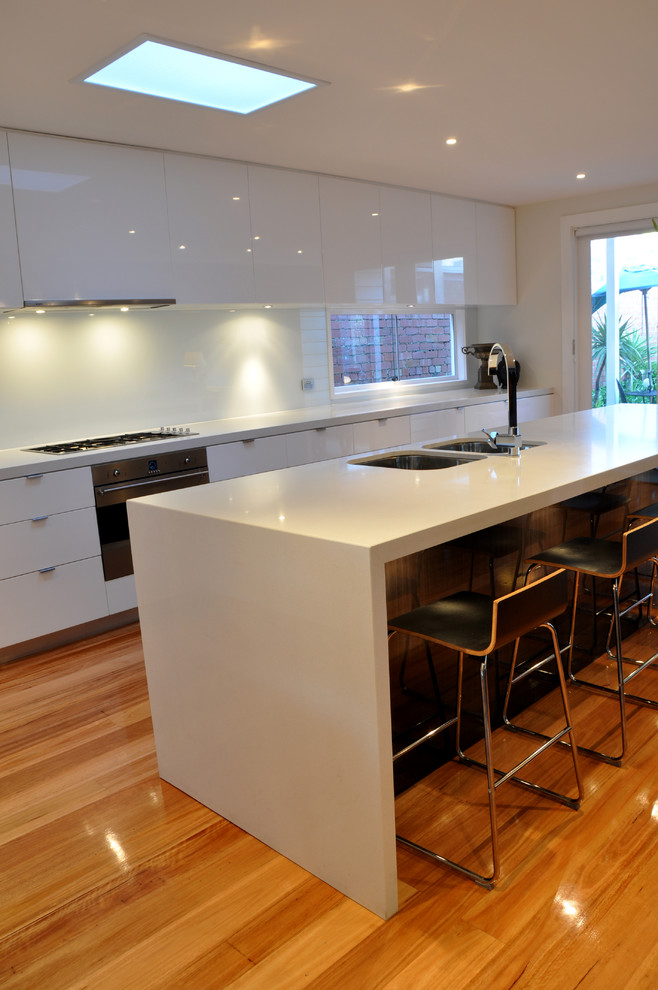 Design ideas for a mid-sized modern l-shaped open plan kitchen in Geelong with an undermount sink, flat-panel cabinets, white cabinets, quartz benchtops, white splashback, glass sheet splashback, stainless steel appliances, light hardwood floors and with island.