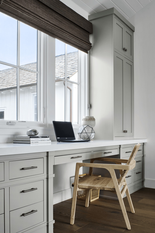 Modelo de despacho blanco marinero de tamaño medio sin chimenea con paredes blancas, suelo de madera en tonos medios, escritorio empotrado, suelo marrón y machihembrado