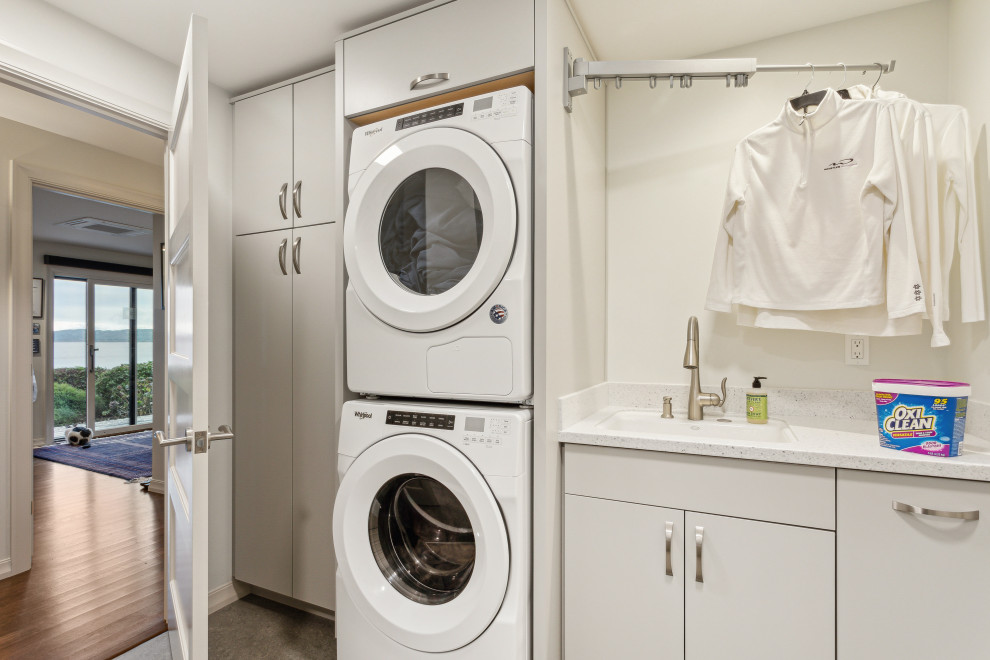 Design ideas for a mid-sized contemporary galley laundry room in Seattle with an undermount sink, flat-panel cabinets, grey cabinets, solid surface benchtops, white splashback, white walls, linoleum floors, a stacked washer and dryer, grey floor and white benchtop.