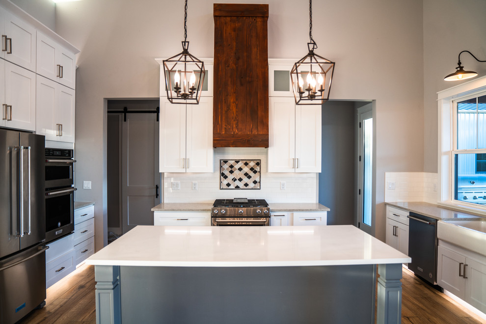 This is an example of a large country u-shaped eat-in kitchen in Austin with a farmhouse sink, recessed-panel cabinets, white cabinets, quartz benchtops, white splashback, subway tile splashback, stainless steel appliances, medium hardwood floors, with island, brown floor and white benchtop.
