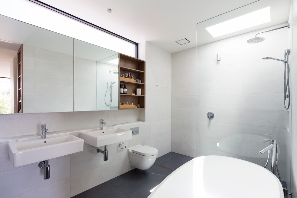 This is an example of a mid-sized contemporary bathroom in Sydney with medium wood cabinets, a freestanding tub, a wall-mount toilet, white tile, ceramic tile, white walls and marble benchtops.