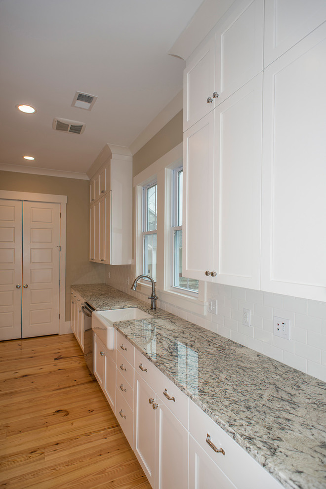 This is an example of a contemporary eat-in kitchen in Charleston with a farmhouse sink, flat-panel cabinets, white cabinets, granite benchtops, white splashback, ceramic splashback, stainless steel appliances, light hardwood floors and with island.