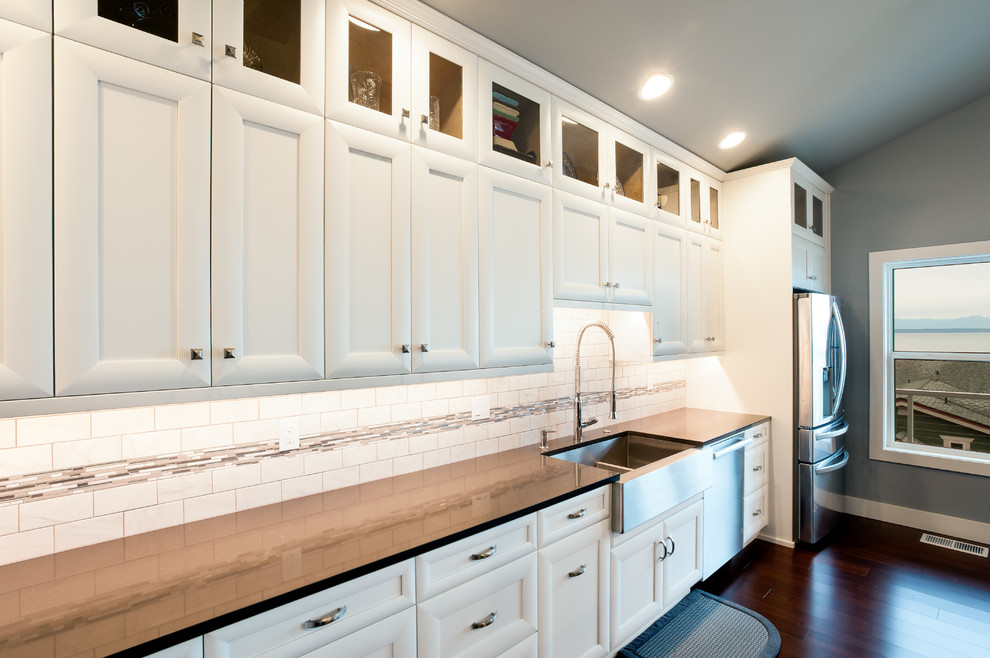 Mid-sized arts and crafts l-shaped open plan kitchen in Seattle with a farmhouse sink, shaker cabinets, white cabinets, quartzite benchtops, white splashback, porcelain splashback, stainless steel appliances, dark hardwood floors, with island, brown floor and white benchtop.