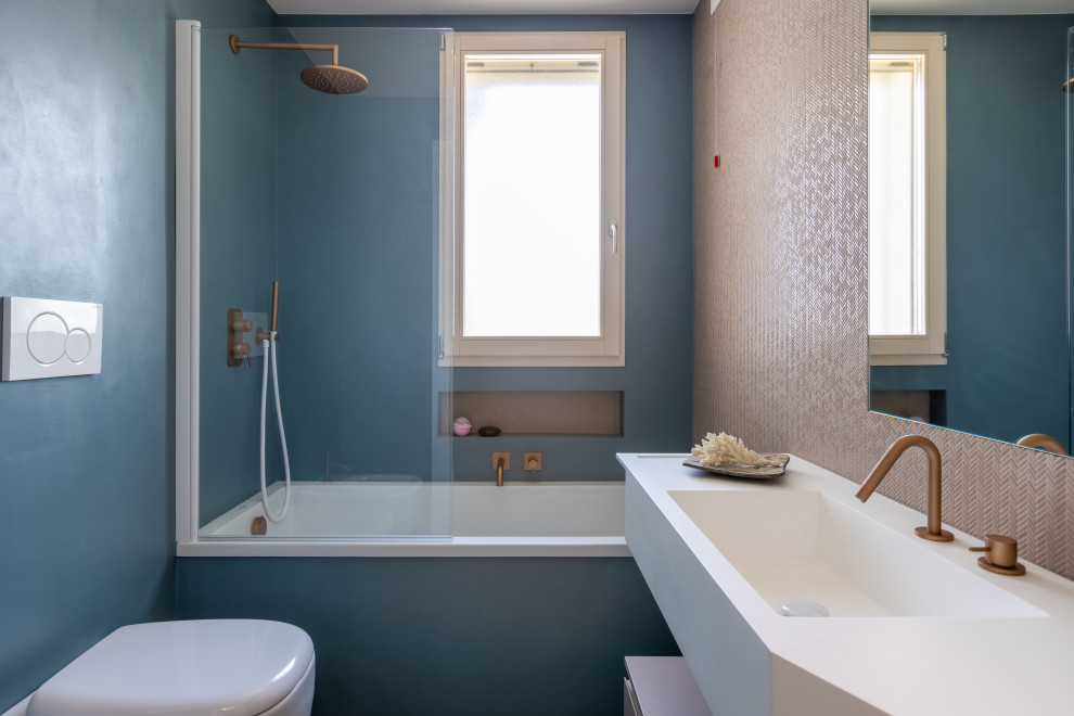 This is an example of a small contemporary ensuite bathroom in Rome with blue cabinets, a built-in bath, a shower/bath combination, a two-piece toilet, pink tiles, mosaic tiles, blue walls, concrete flooring, an integrated sink, solid surface worktops, blue floors, a hinged door, white worktops, a single sink and a floating vanity unit.