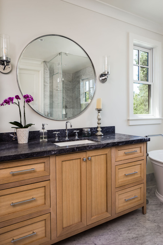 This is an example of a large transitional master bathroom in San Francisco with shaker cabinets, light wood cabinets, a freestanding tub, a curbless shower, a one-piece toilet, white tile, marble, white walls, marble floors, an undermount sink, marble benchtops, white floor, a hinged shower door, black benchtops, a niche, a double vanity and a built-in vanity.
