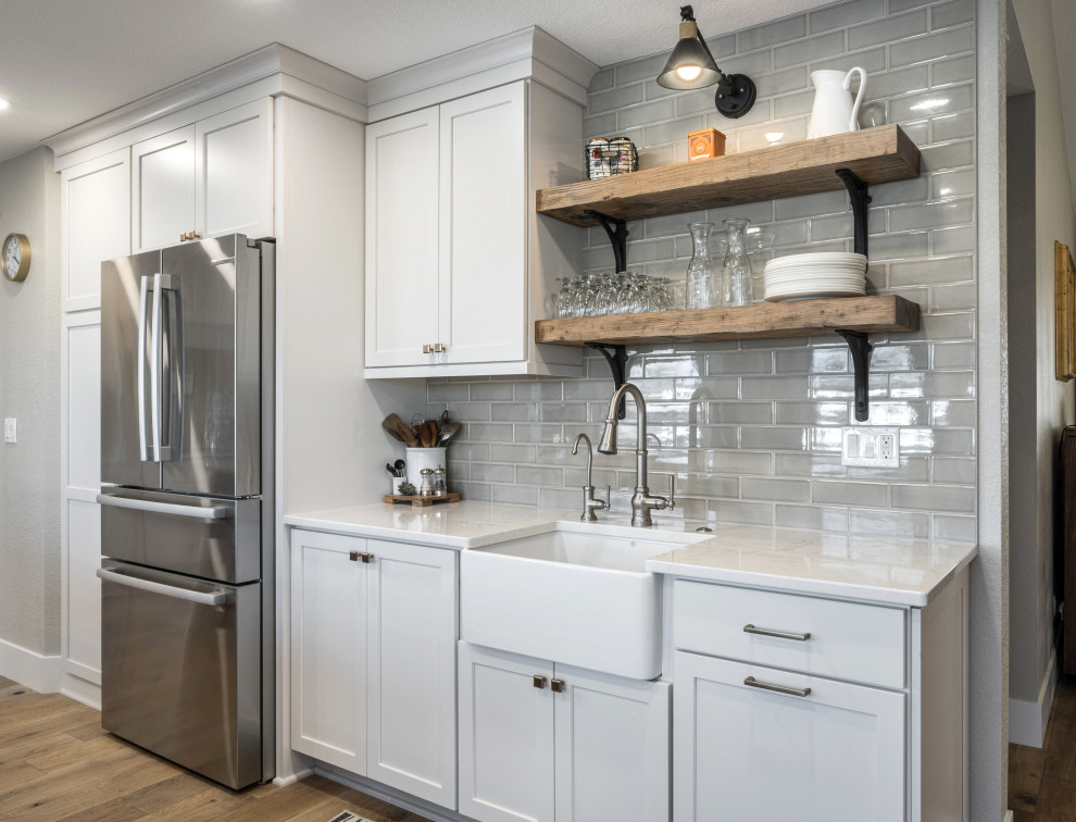 Inspiration for a large country l-shaped open plan kitchen in Boise with a belfast sink, shaker cabinets, white cabinets, engineered stone countertops, grey splashback, porcelain splashback, stainless steel appliances, medium hardwood flooring, an island, brown floors and white worktops.