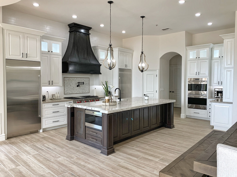 Large transitional l-shaped open plan kitchen in Phoenix with a farmhouse sink, raised-panel cabinets, white cabinets, quartz benchtops, white splashback, ceramic splashback, stainless steel appliances, cement tiles, with island, grey floor and beige benchtop.