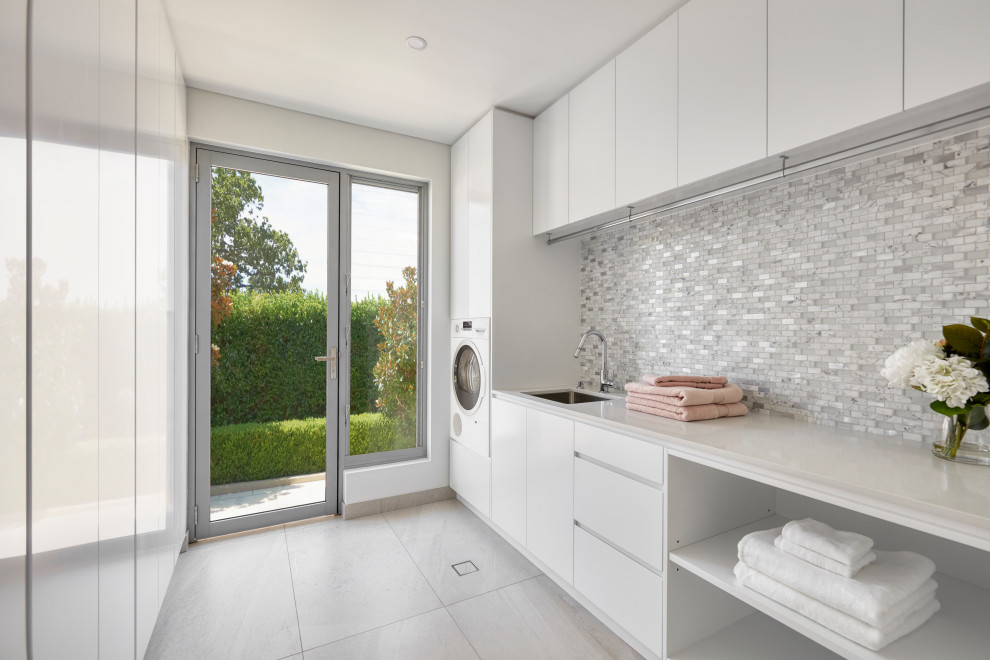 Design ideas for a mid-sized contemporary l-shaped dedicated laundry room in Sydney with an undermount sink, flat-panel cabinets, white cabinets, granite benchtops, grey splashback, mosaic tile splashback, white walls, porcelain floors, an integrated washer and dryer, grey floor and white benchtop.