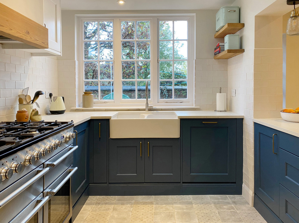 This is an example of a medium sized contemporary u-shaped open plan kitchen in Other with a belfast sink, shaker cabinets, blue cabinets, granite worktops, white splashback, ceramic splashback, stainless steel appliances, cement flooring, a breakfast bar, grey floors, grey worktops and a chimney breast.
