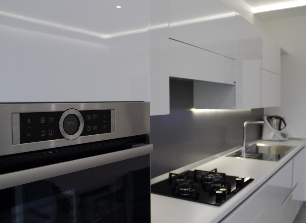 This is an example of a mid-sized contemporary single-wall open plan kitchen in Bari with a drop-in sink, flat-panel cabinets, white cabinets, solid surface benchtops, grey splashback, porcelain splashback, stainless steel appliances, porcelain floors, no island, grey floor and white benchtop.