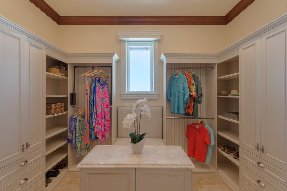 This is an example of a large beach style gender-neutral walk-in wardrobe in Miami with beaded inset cabinets, white cabinets, travertine floors and beige floor.