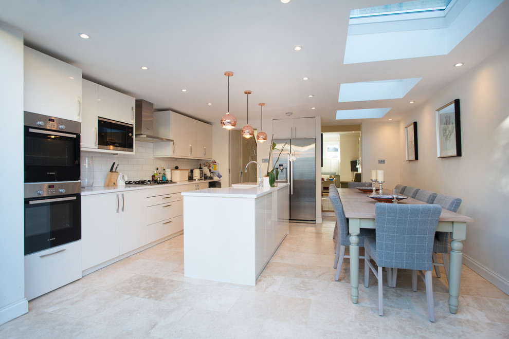 Photo of a mid-sized modern single-wall eat-in kitchen in London with a farmhouse sink, flat-panel cabinets, white cabinets, white splashback, stainless steel appliances, with island, beige floor and white benchtop.