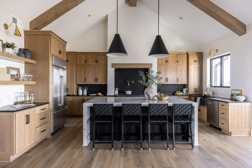 This is an example of a transitional u-shaped kitchen in Sydney with a farmhouse sink, shaker cabinets, light wood cabinets, black splashback, stone slab splashback, stainless steel appliances, medium hardwood floors, with island, brown floor, black benchtop, exposed beam and vaulted.
