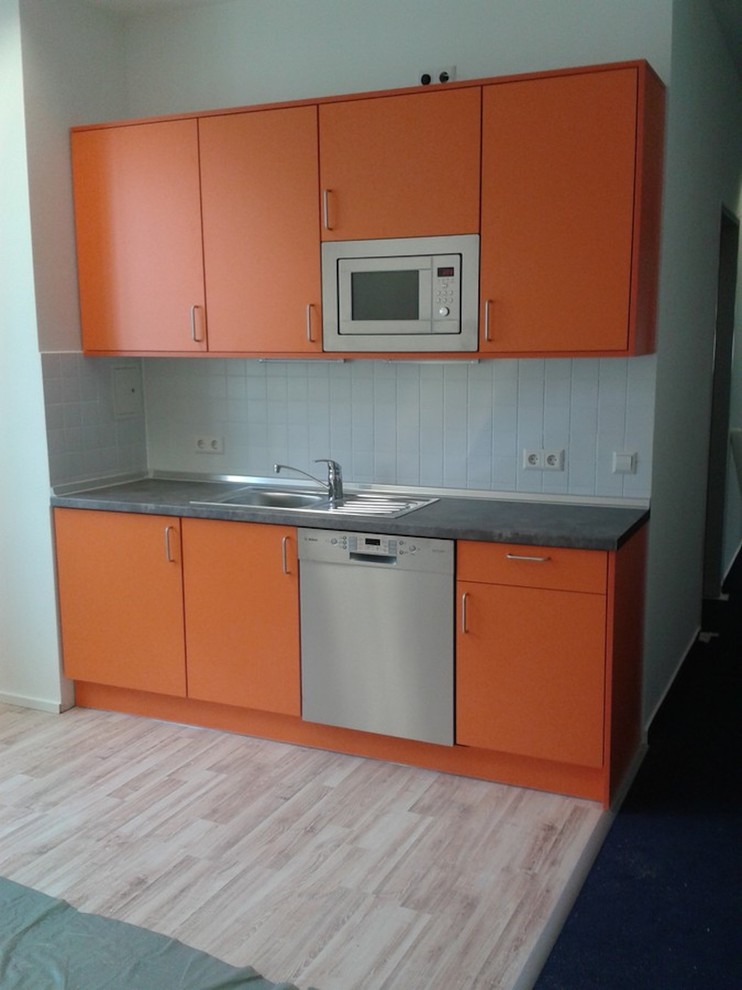 This is an example of a small contemporary single-wall open plan kitchen in Berlin with an integrated sink, flat-panel cabinets, orange cabinets, wood benchtops, white splashback, stainless steel appliances, laminate floors, no island and brown floor.