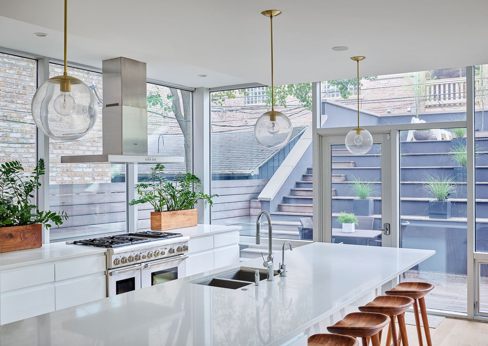 Photo of a contemporary kitchen in Chicago with a double-bowl sink, flat-panel cabinets, white cabinets, window splashback, stainless steel appliances, light hardwood floors, with island, brown floor and white benchtop.