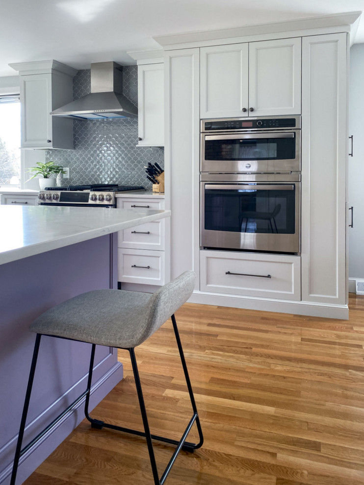 This is an example of a traditional l-shaped kitchen in Boston with a belfast sink, shaker cabinets, purple cabinets, engineered stone countertops, grey splashback, ceramic splashback, stainless steel appliances, light hardwood flooring, an island and white worktops.