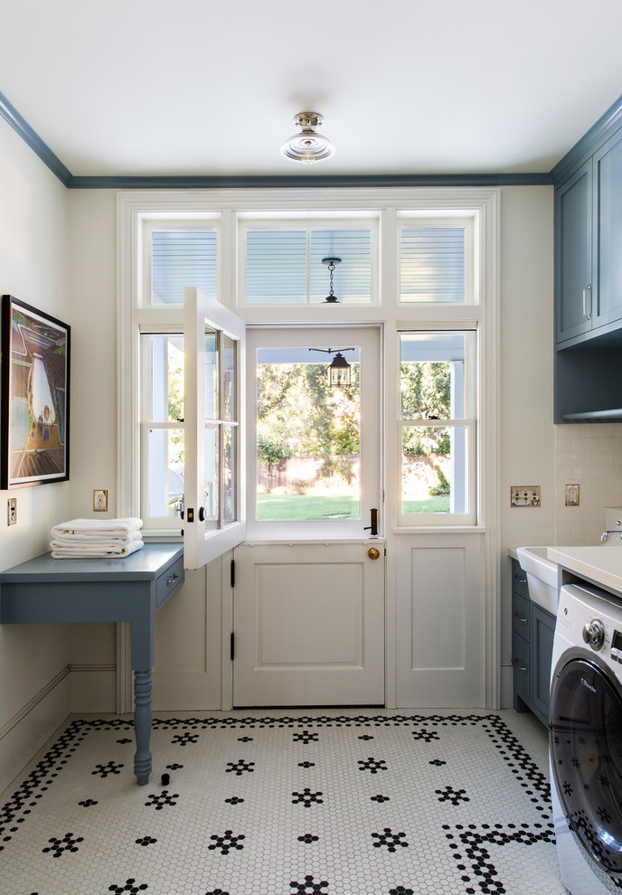 This is an example of a large traditional single-wall laundry room in San Francisco with a farmhouse sink, shaker cabinets, blue cabinets, white walls, a side-by-side washer and dryer, quartzite benchtops, ceramic floors, multi-coloured floor and white benchtop.