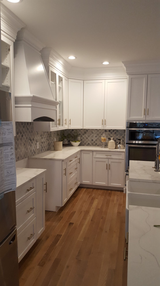Large transitional u-shaped light wood floor and brown floor eat-in kitchen photo in DC Metro with a farmhouse sink, recessed-panel cabinets, white cabinets, quartzite countertops, gray backsplash, marble backsplash, stainless steel appliances, an island and white countertops