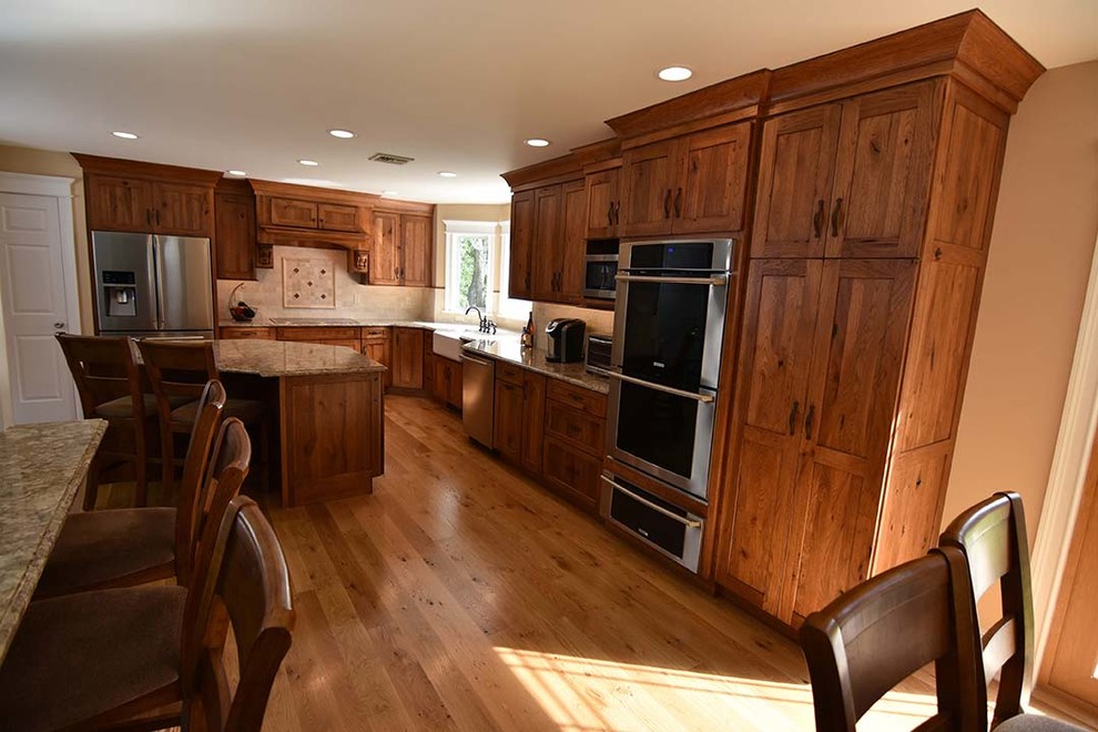 Inspiration for a country l-shaped open plan kitchen in Orange County with a farmhouse sink, shaker cabinets, medium wood cabinets, granite benchtops, beige splashback, ceramic splashback, stainless steel appliances, light hardwood floors, with island and brown floor.