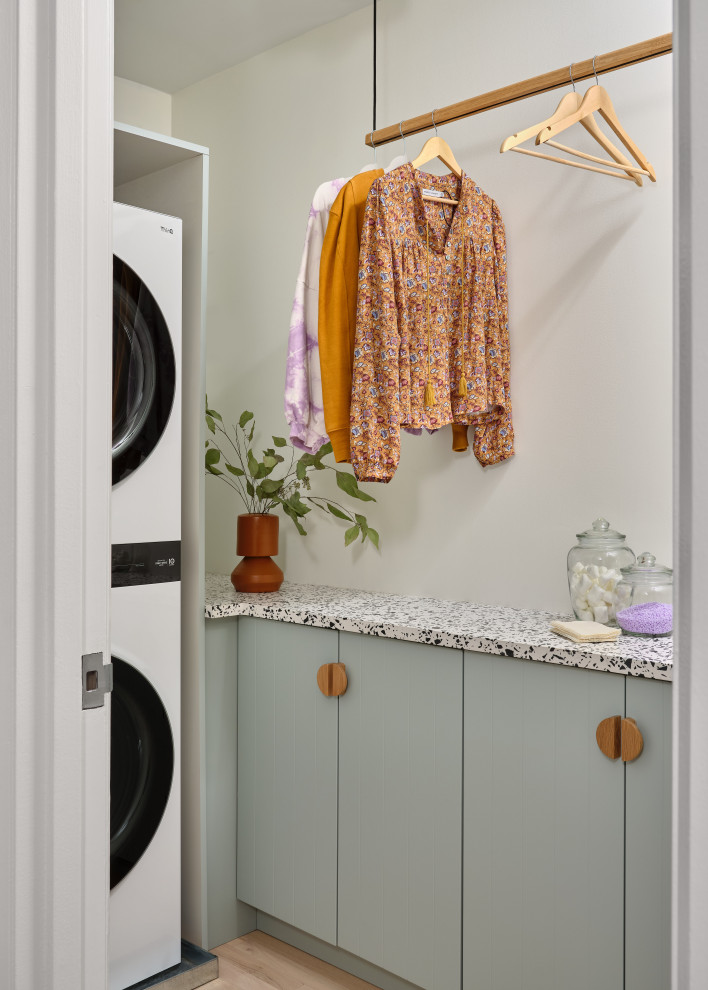 Photo of a contemporary l-shaped utility room in New York with flat-panel cabinets, green cabinets, beige walls, light hardwood flooring, a stacked washer and dryer, beige floors and multicoloured worktops.