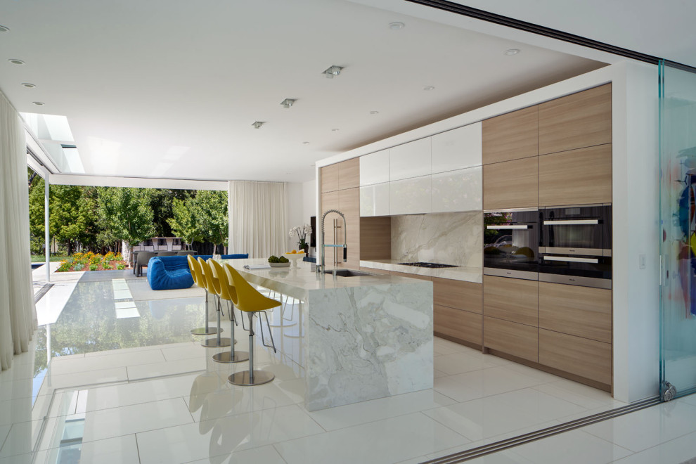 This is an example of a large contemporary single-wall open plan kitchen in San Francisco with an undermount sink, flat-panel cabinets, medium wood cabinets, marble benchtops, white splashback, marble splashback, black appliances, ceramic floors, with island, white floor and white benchtop.