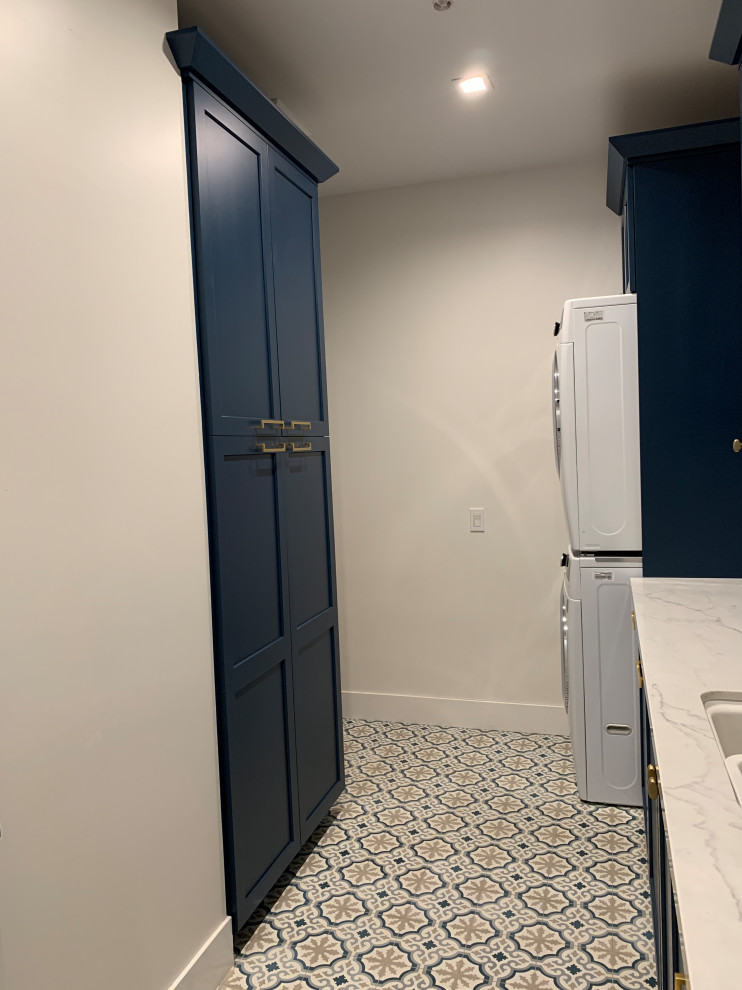 Photo of a mid-sized beach style galley dedicated laundry room in Orange County with a double-bowl sink, shaker cabinets, blue cabinets, quartz benchtops, white splashback, ceramic splashback, white walls, ceramic floors, a stacked washer and dryer, multi-coloured floor and white benchtop.