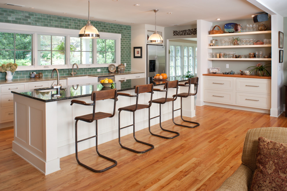 Example of a large transitional medium tone wood floor and brown floor open concept kitchen design with a farmhouse sink, shaker cabinets, white cabinets, green backsplash, subway tile backsplash, stainless steel appliances and an island