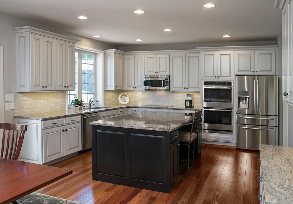 This is an example of a mid-sized traditional l-shaped eat-in kitchen in Philadelphia with an undermount sink, raised-panel cabinets, white cabinets, granite benchtops, white splashback, ceramic splashback, stainless steel appliances, medium hardwood floors, with island and brown floor.